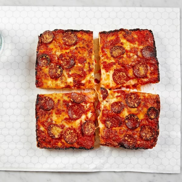 Four square slices of Detroit-style pepperoni pizza with crispy edges and melted cheese on white paper.