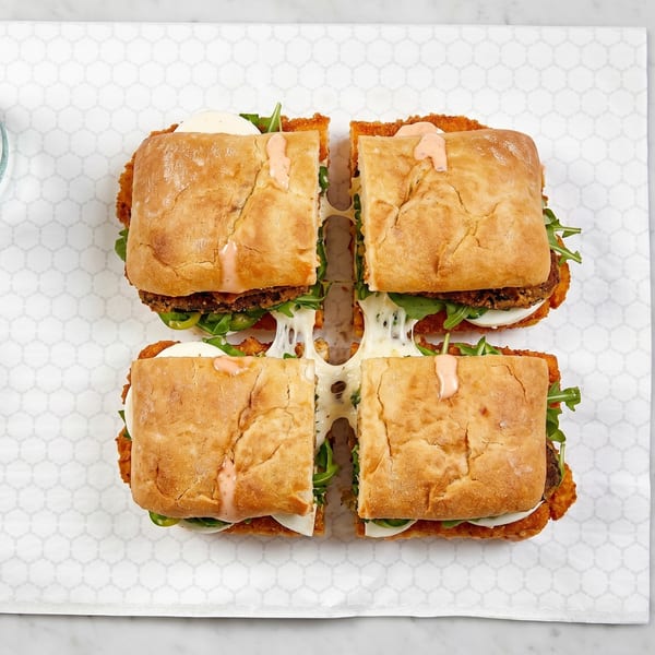 Four halves of a crispy chicken sandwich with melted cheese, arugula, and sauce on ciabatta bread.