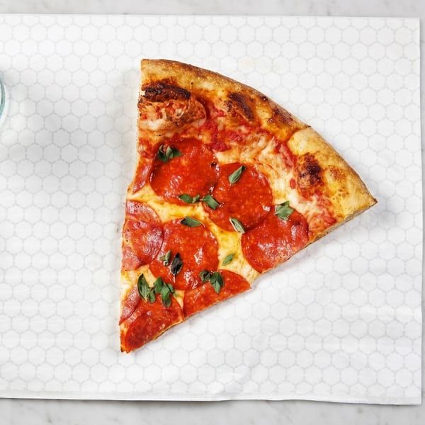 A single slice of pepperoni pizza with fresh basil on white patterned paper.