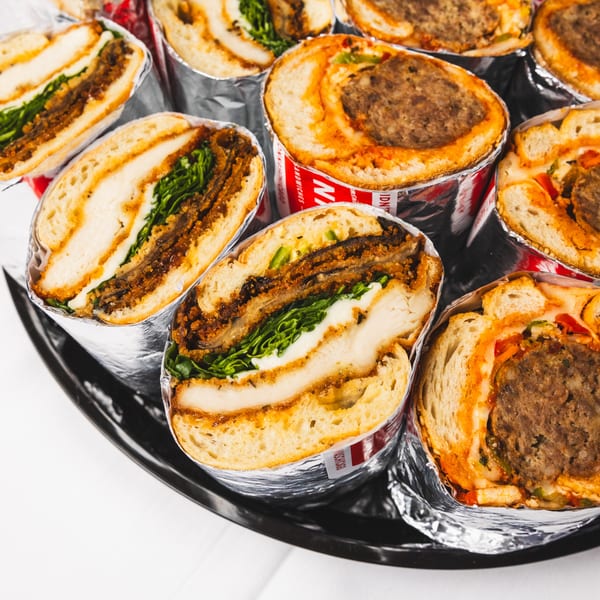A black tray filled with foil-wrapped sandwiches, some with fried chicken and greens, others with meatballs and sauce.