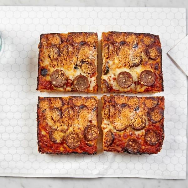 Four square slices of Detroit-style pizza with pepperoni, sausage, and crispy cheese on a white paper.